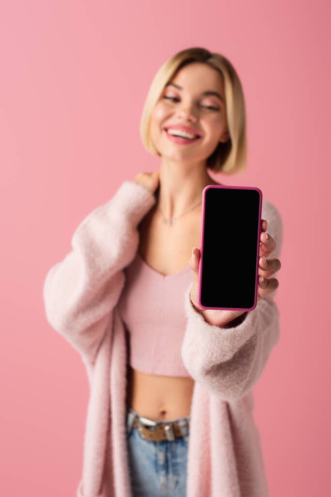 verschwommen und glücklich junge Frau in weicher Strickjacke hält Smartphone mit leerem Bildschirm isoliert auf rosa  - Foto, Bild