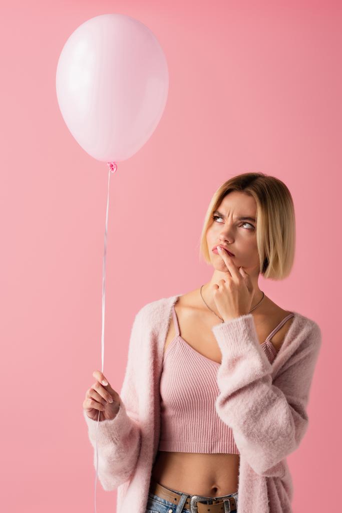 σκεπτική νεαρή γυναίκα σε ζακέτα κρατώντας μπαλόνι απομονωμένο σε ροζ - Φωτογραφία, εικόνα