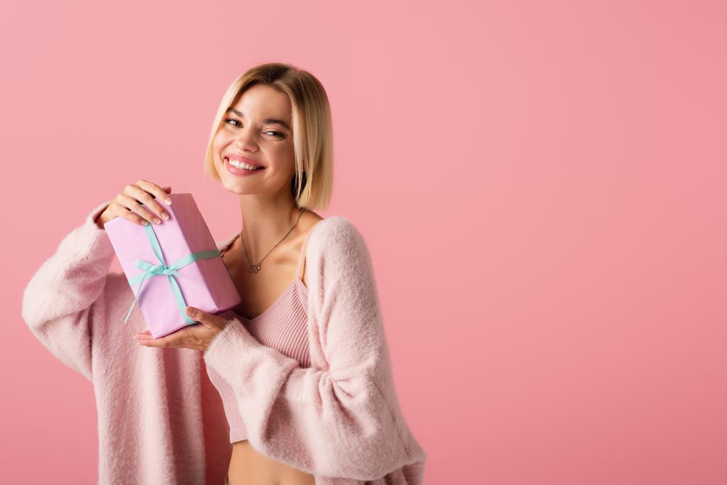 Χαρούμενη νεαρή γυναίκα σε ζακέτα κρατώντας τυλιγμένο κουτί δώρου απομονωμένο σε ροζ - Φωτογραφία, εικόνα
