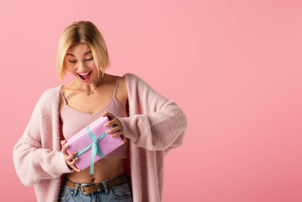 amazed young woman in cardigan holding wrapped gift box isolated on pink - Photo, Image