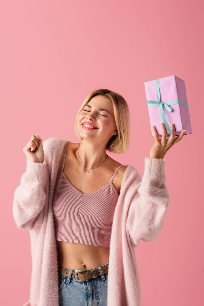 heureuse jeune femme avec les yeux fermés tenant boîte cadeau enveloppé isolé sur rose - Photo, image
