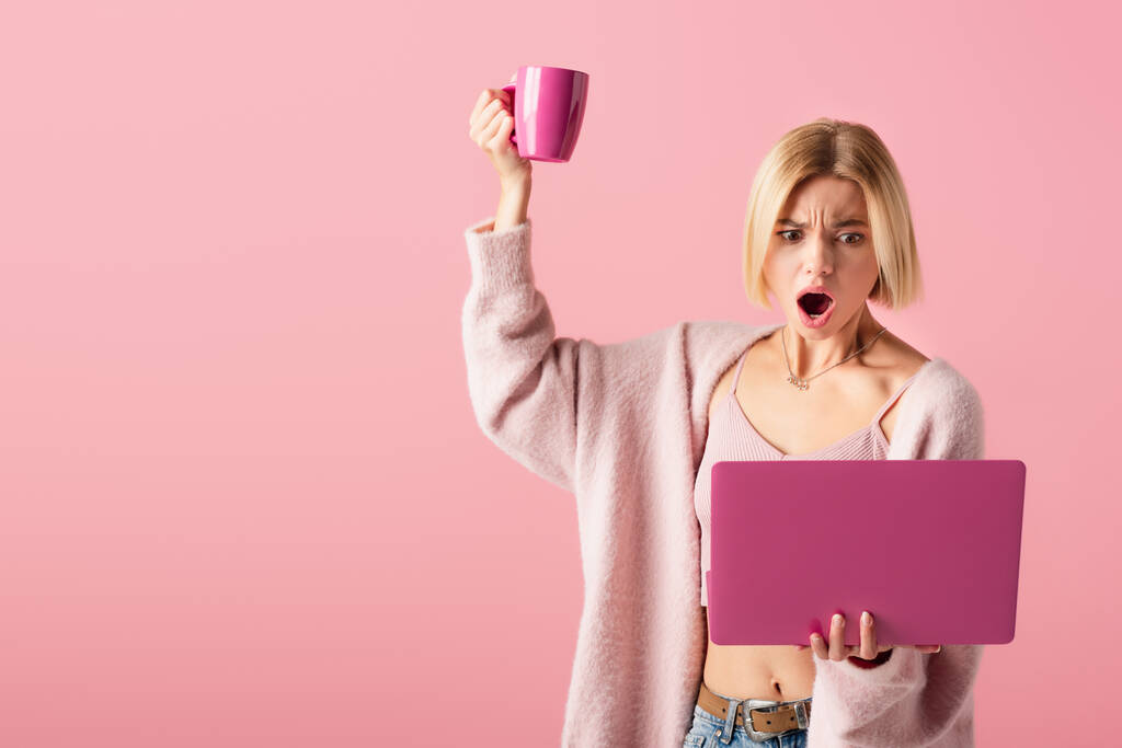 ショックを受けた女性がマグカップとコーヒーとノートパソコンをピンクに隔離し  - 写真・画像