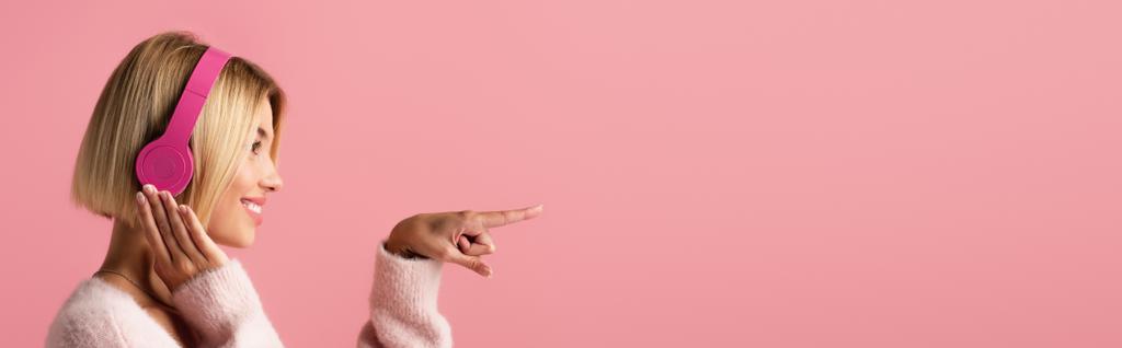 Seitenansicht einer fröhlichen Frau, die Musik in drahtlosen Kopfhörern hört und mit dem Finger isoliert auf rosa zeigt, Banner - Foto, Bild