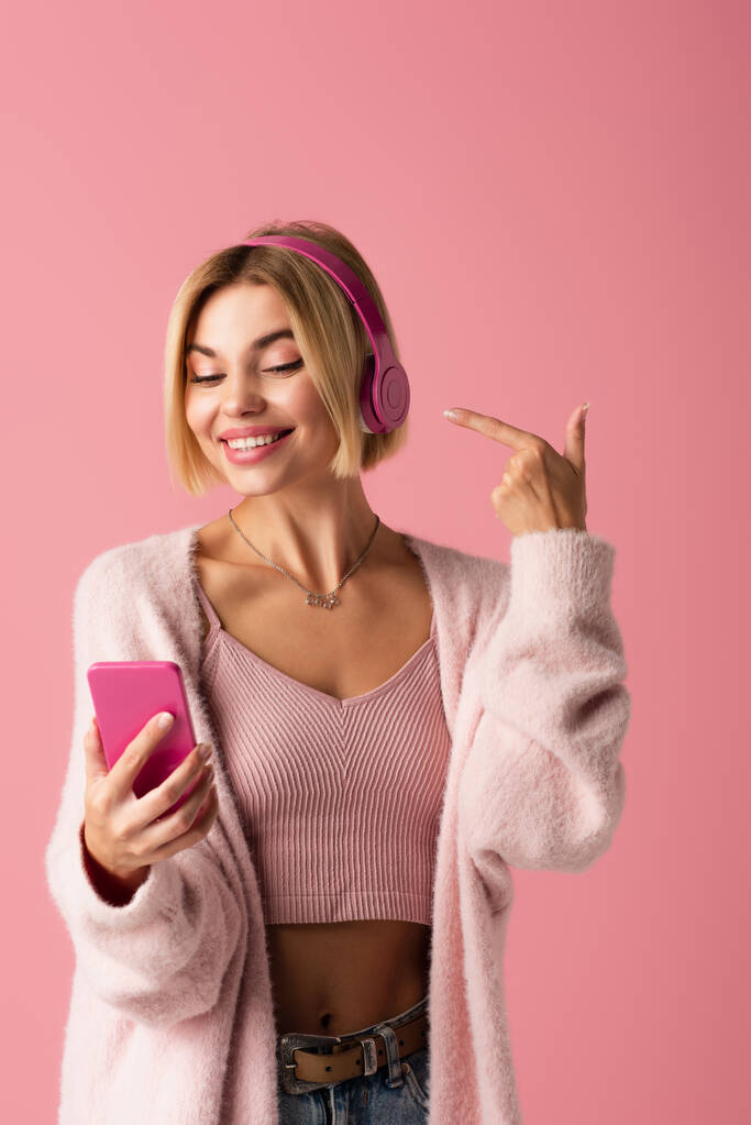 veselá žena ukazuje na bezdrátová sluchátka a drží smartphone izolované na růžové  - Fotografie, Obrázek