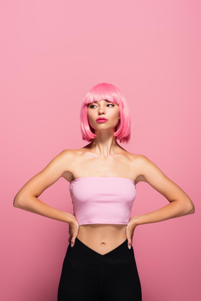 mladý model s ofinou a barevnými vlasy pózující s rukama na bocích izolovaných na růžové - Fotografie, Obrázek