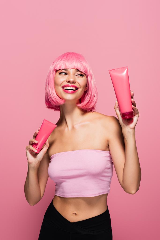 radosna młoda kobieta z kolorowymi włosami i grzywką trzyma rury z balsamem odizolowanym na różowo  - Zdjęcie, obraz