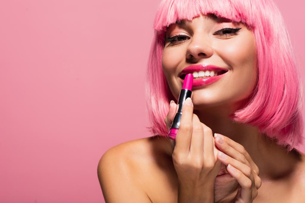 mujer joven feliz con el pelo de color y el hombro desnudo aplicando lápiz labial aislado en rosa  - Foto, Imagen