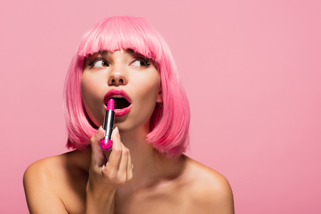 giovane donna con i capelli colorati e le spalle nude applicando rossetto e distogliendo lo sguardo isolato sul rosa  - Foto, immagini