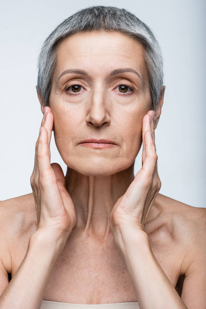 Frau mittleren Alters berührt Gesicht mit Falten isoliert auf grau - Foto, Bild