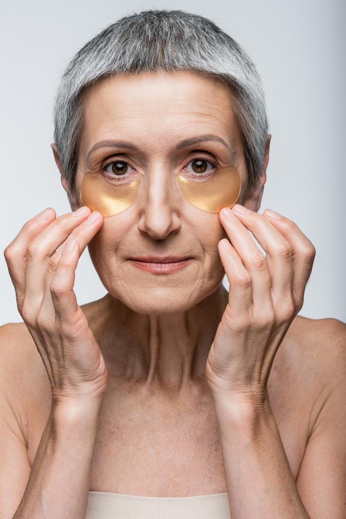 vrouw van middelbare leeftijd aanbrengen van collageen ooglapjes en kijken naar camera geïsoleerd op grijs - Foto, afbeelding