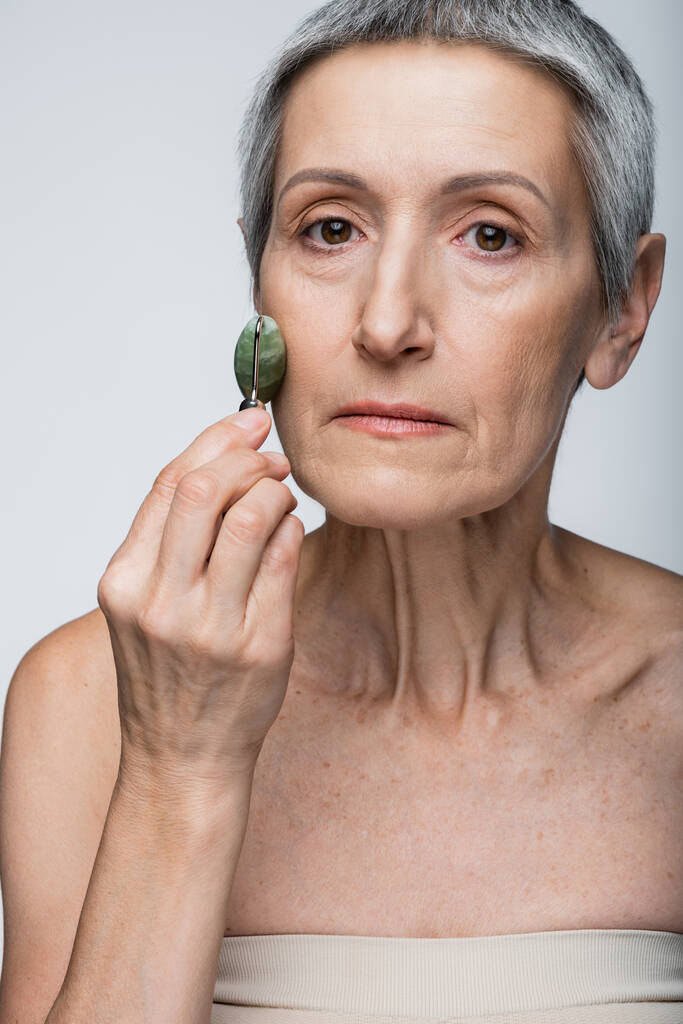 femme d'âge moyen massant la joue avec rouleau de jade isolé sur gris  - Photo, image