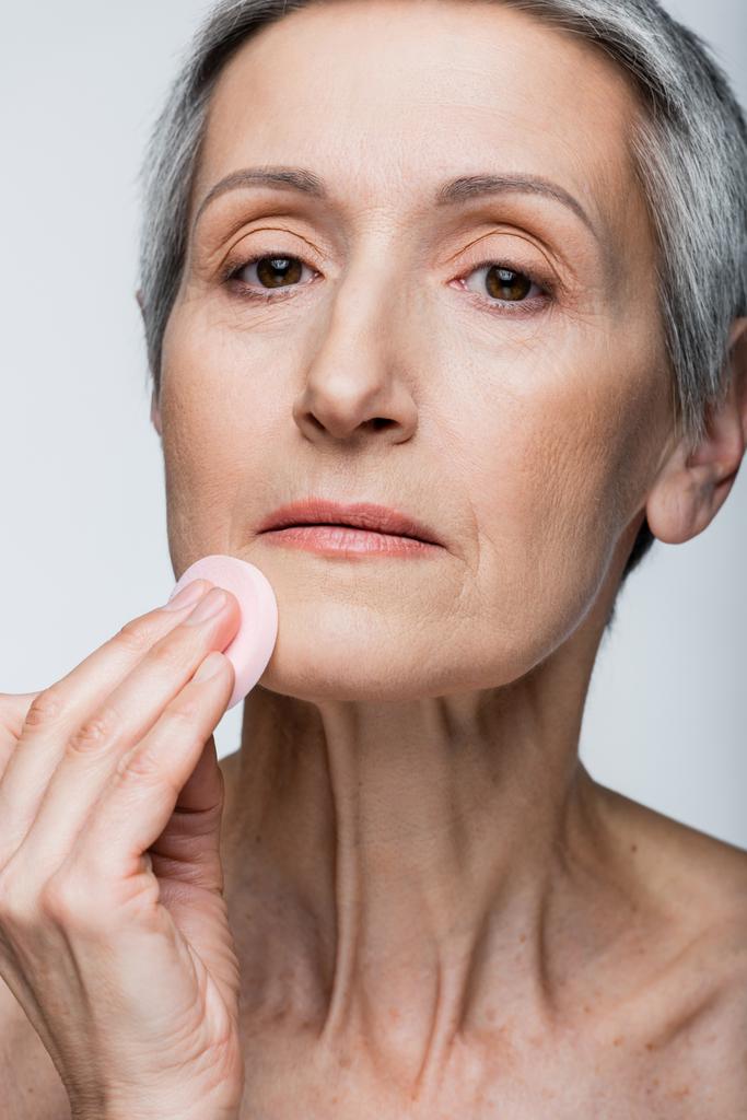 close up of mature woman with grey hair holding beauty sponge and applying makeup foundation isolated on grey  - Photo, Image
