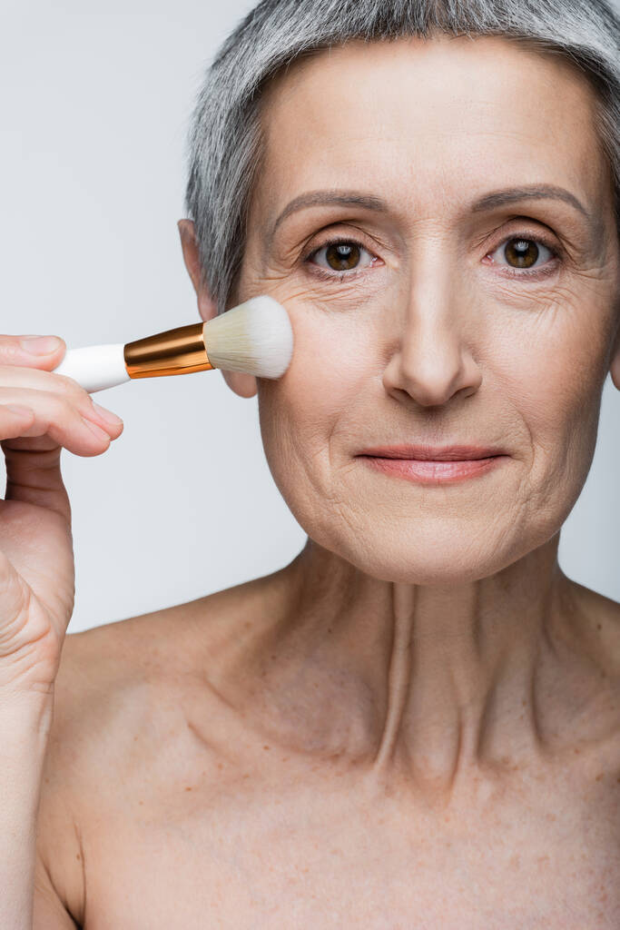middle aged woman with bare shoulders applying face powder on cheek with cosmetic brush isolated on grey - Photo, Image