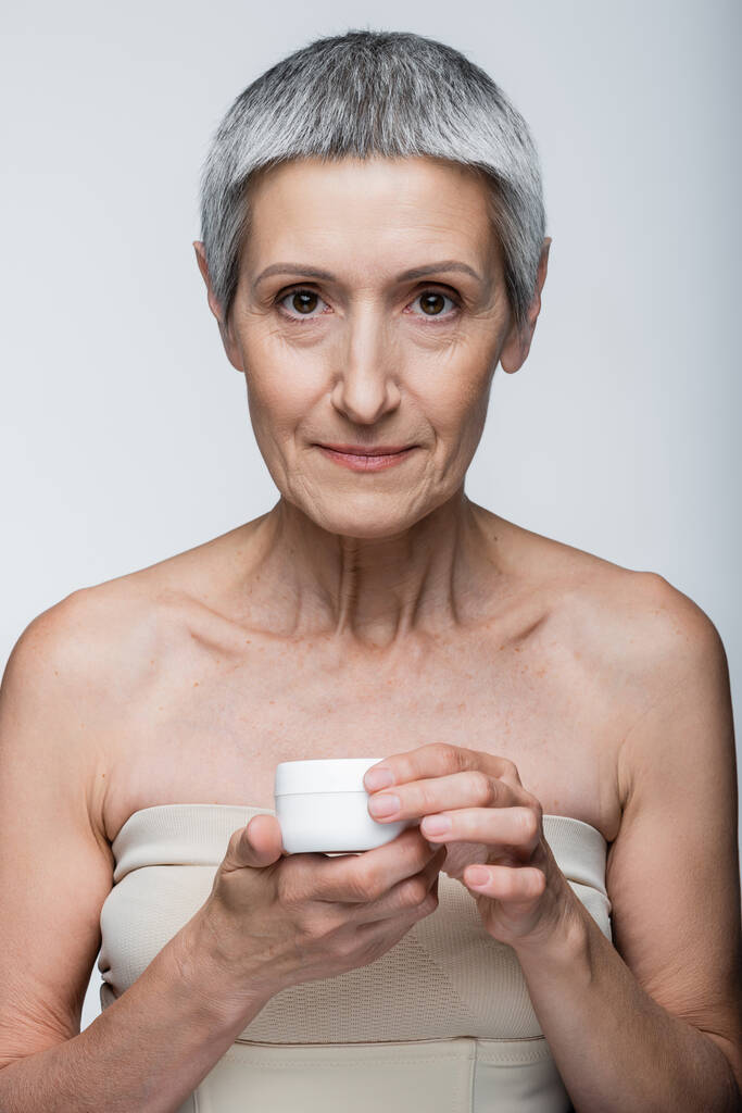 volwassen vrouw met blote schouders houden container met gezicht crème geïsoleerd op grijs  - Foto, afbeelding