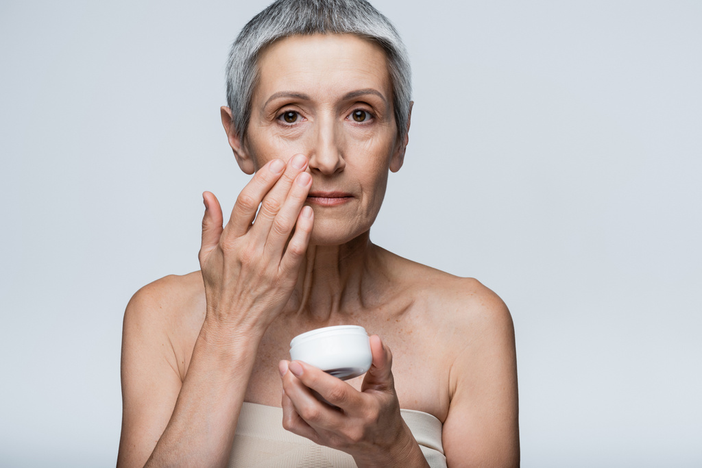 mature woman with grey hair holding container and applying cosmetic cream isolated on grey  - Photo, Image