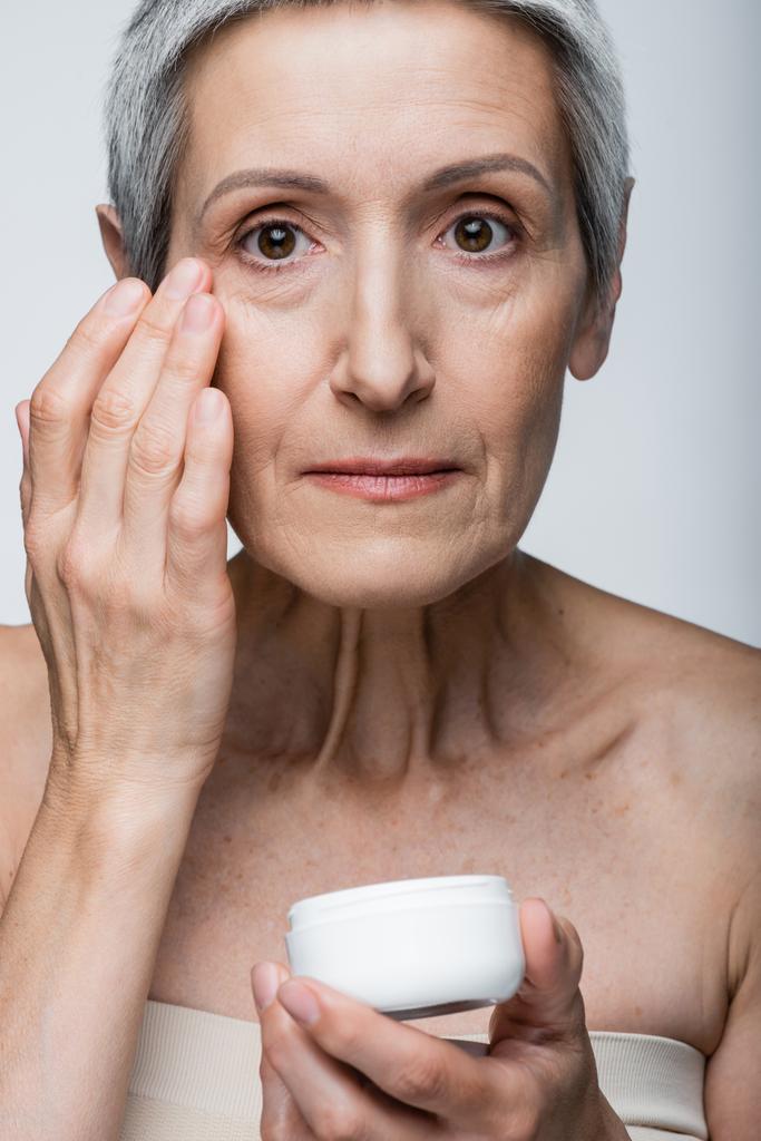 volwassen vrouw met grijs haar aanbrengen anti-rimpel crème geïsoleerd op grijs  - Foto, afbeelding