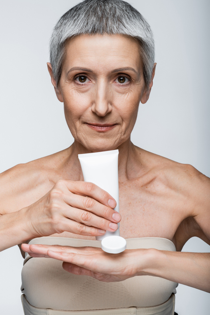 donna di mezza età con tubo di tenuta dei capelli grigi e applicare lozione a mano isolata su grigio  - Foto, immagini