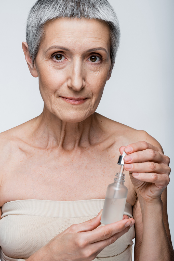 dojrzała kobieta z siwymi włosami trzymająca butelkę i pipetą z serum nawilżającym wyizolowanym na szaro - Zdjęcie, obraz