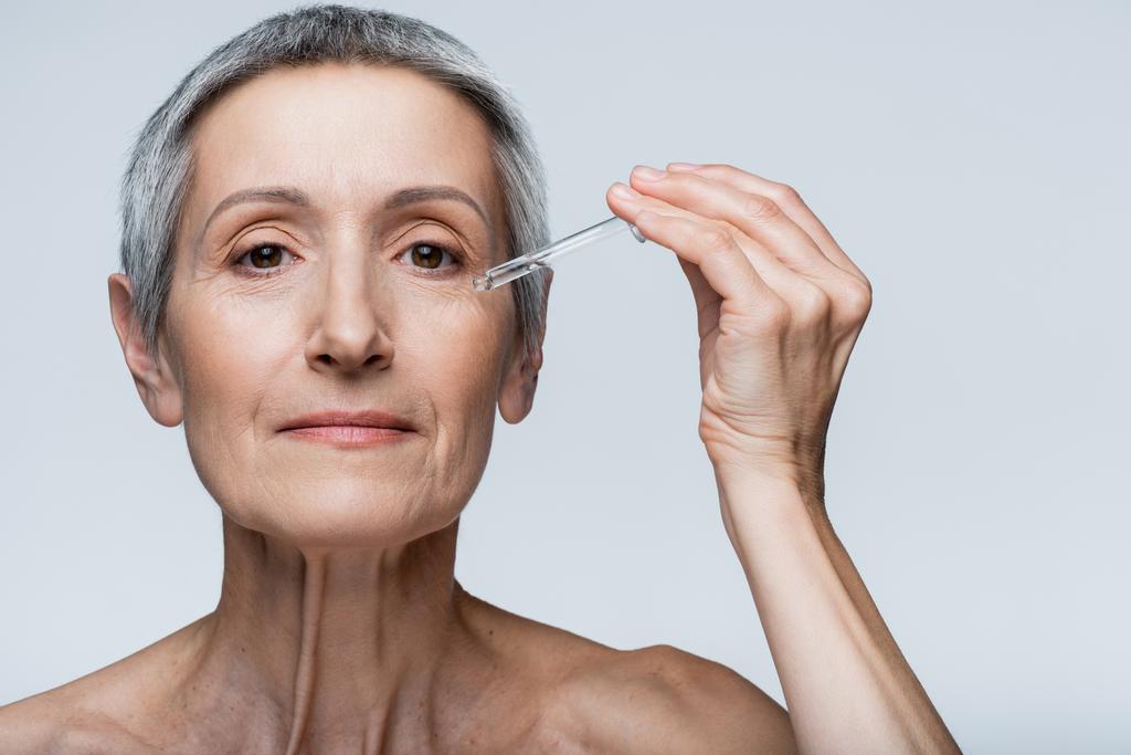 middle aged woman with grey hair holding pipette with serum isolated on grey - Photo, Image