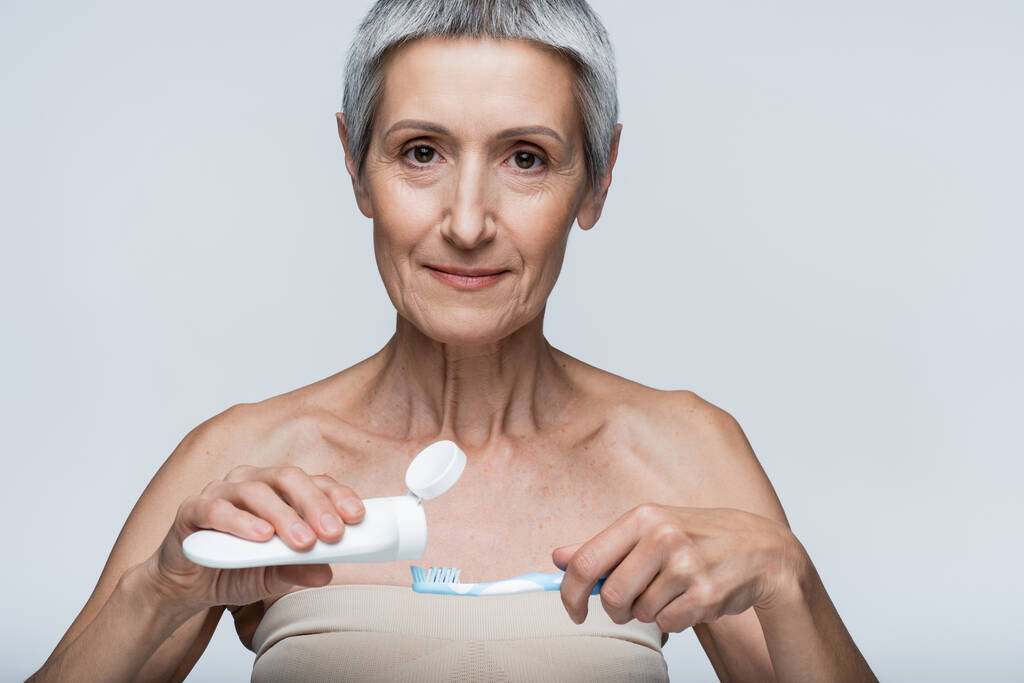 жінка середнього віку тримає зубну щітку і трубку з зубною пастою ізольовано на сірому
  - Фото, зображення