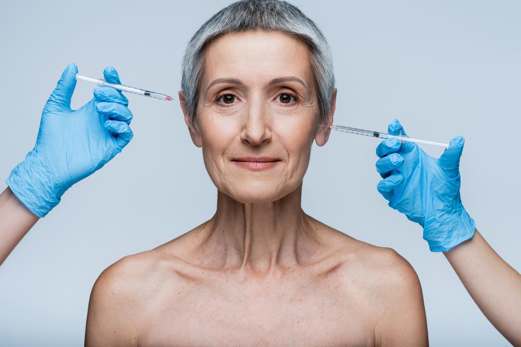 beauticians in latex gloves holding syringe with beauty injection near smiling middle aged client isolated on grey - Photo, Image