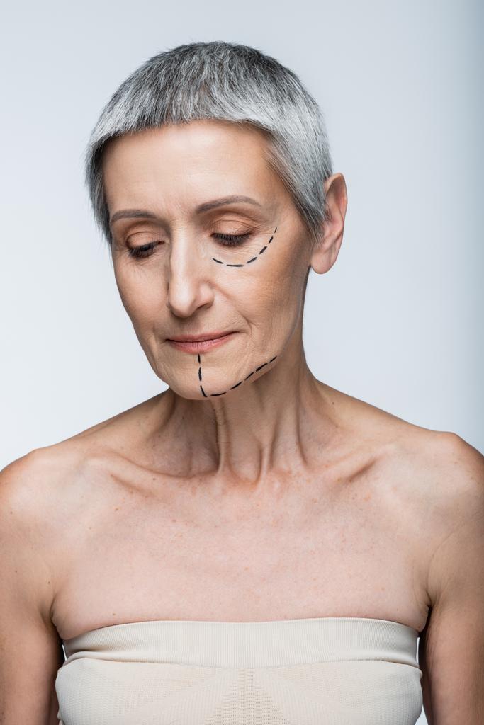 volwassen vrouw met blote schouders en gemarkeerde lijnen op gezicht naar beneden kijken geïsoleerd op grijs - Foto, afbeelding