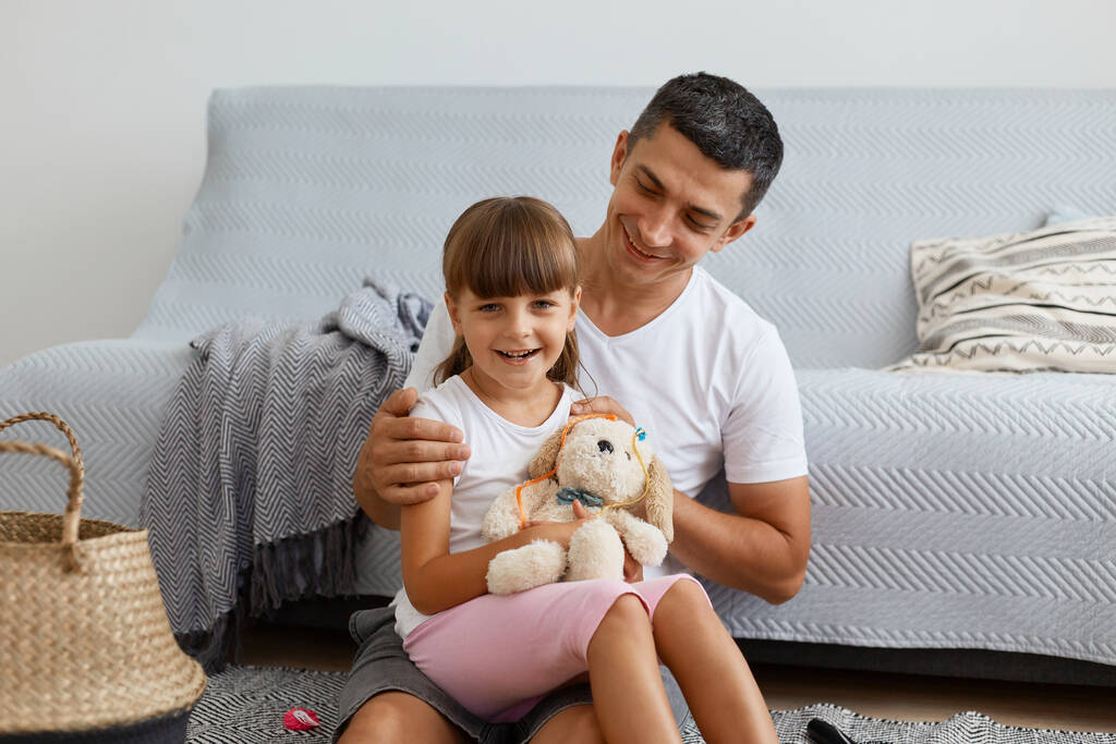 Indoor shot of smiling brunette man wearing white casual style T-shirt and jeans short sitting on floor with his daughter, kid holding toy, family expressing positive emotions. - Photo, Image