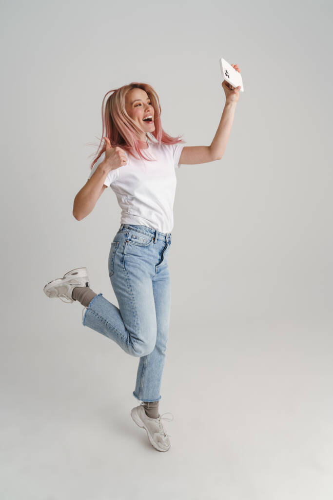 Boldog fiatal nő alkalmi viselet jumping csinál szelfi tartó mobiltelefon felett szürke háttér, teljes hossz - Fotó, kép