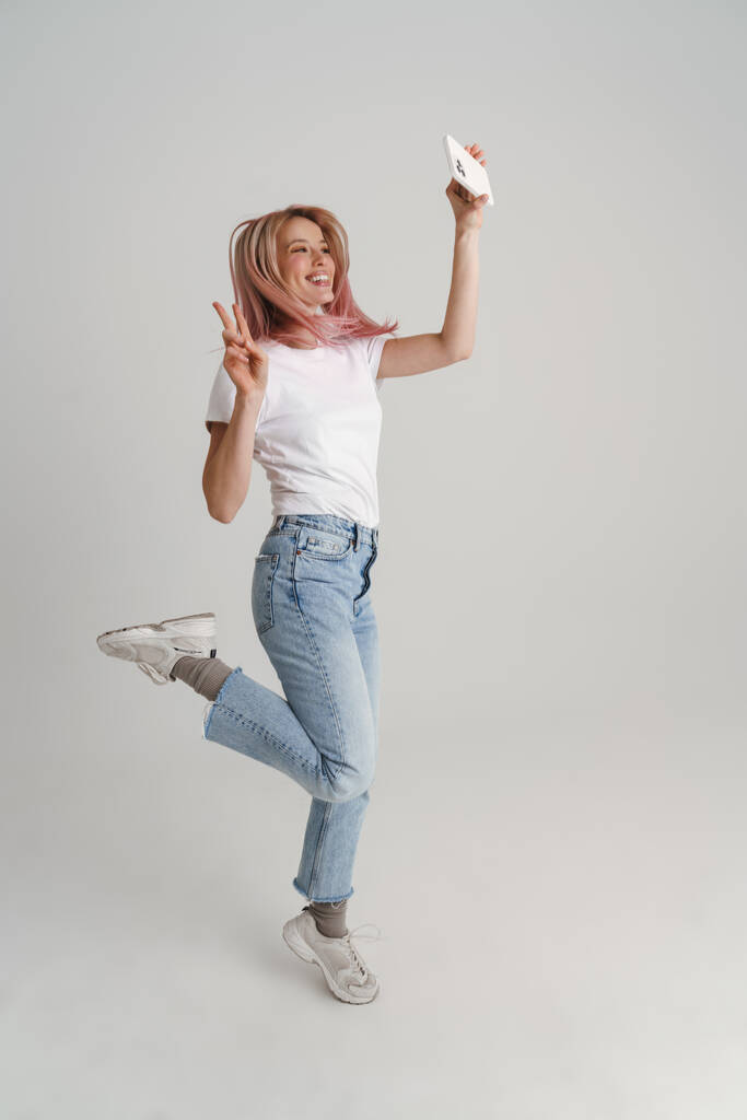 Szczęśliwa młoda kobieta w casual wear jumping biorąc selfie trzyma telefon komórkowy na szarym tle, pełna długość - Zdjęcie, obraz