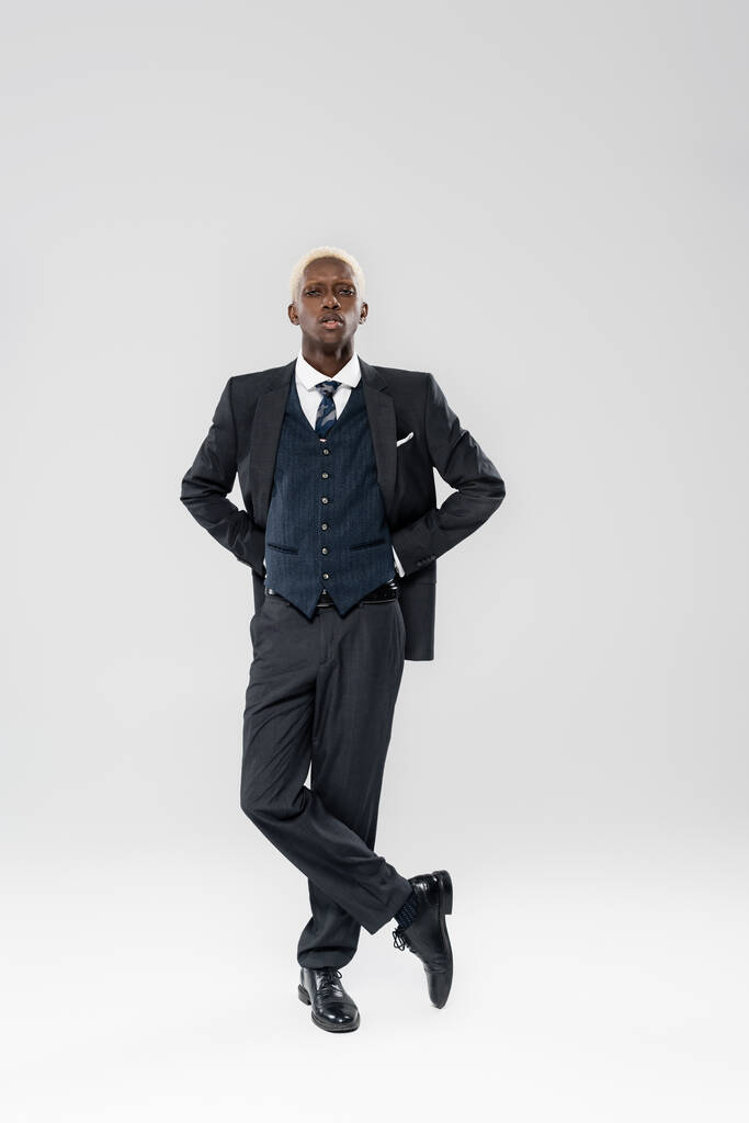 volle Länge der blonde afrikanisch-amerikanische Mann im Anzug stehend mit überkreuzten Beinen auf grau - Foto, Bild