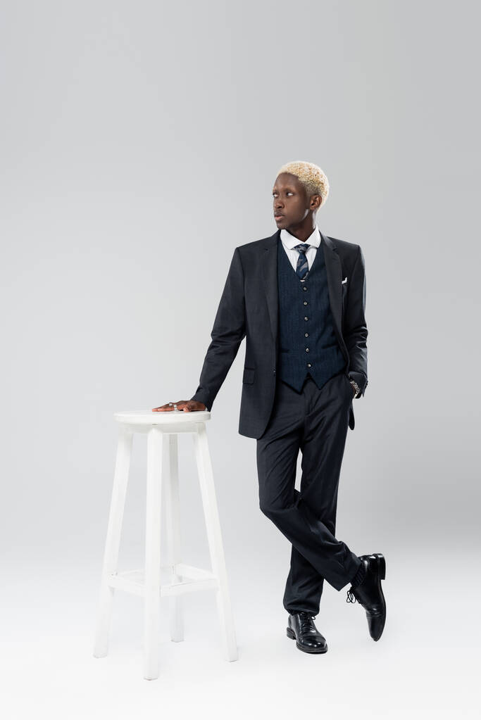 longitud completa de hombre afroamericano rubio en traje de pie con la mano en el bolsillo cerca de la silla blanca en gris - Foto, Imagen
