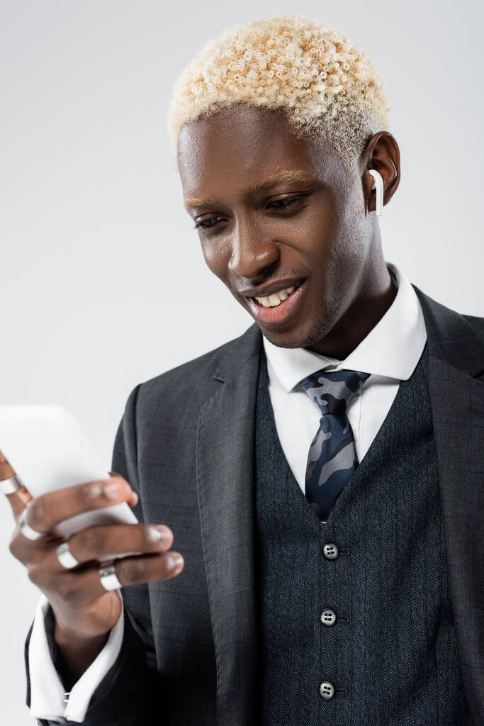 hombre americano africano alegre en auriculares inalámbricos usando teléfono inteligente aislado en gris - Foto, Imagen
