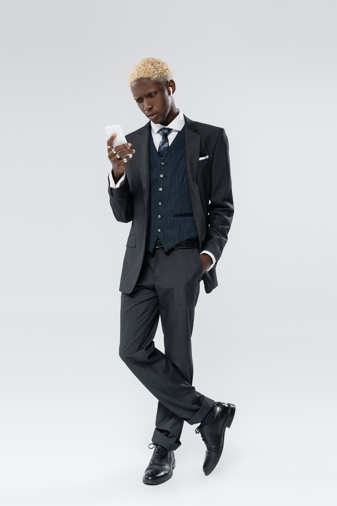 longitud completa de hombre afroamericano rubia en auriculares inalámbricos con teléfono inteligente y posando en gris - Foto, Imagen