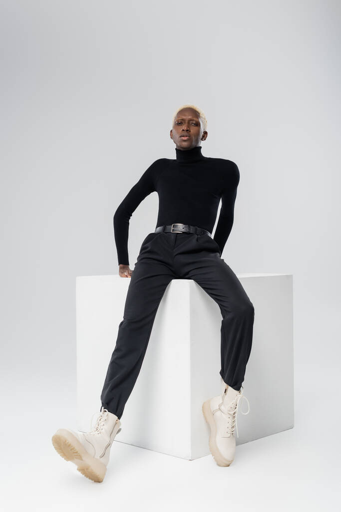 full length of young african american man in turtleneck posing on white cube on grey  - Photo, Image