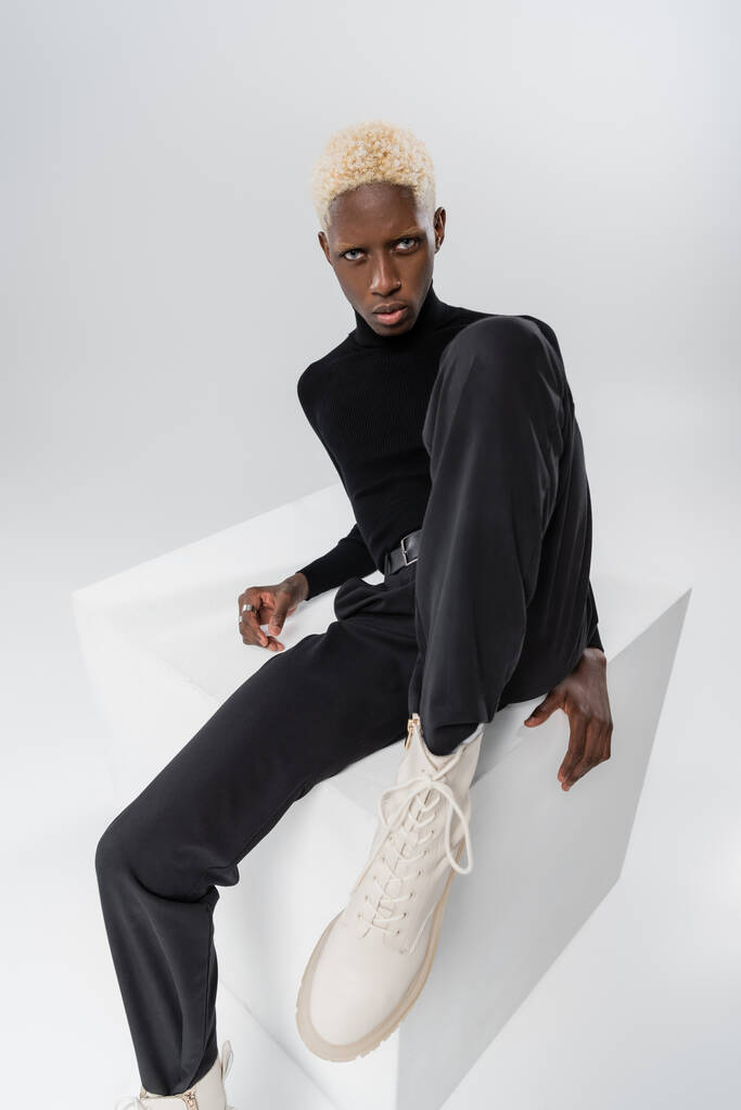 ξανθός αφροαμερικανός νεαρός άνδρας κάθεται σε λευκό κύβο απομονωμένος σε γκρι  - Φωτογραφία, εικόνα