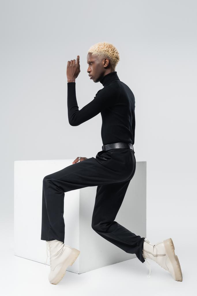 side view of young african american man in stylish outfit sitting on white cube and posing on grey  - Photo, Image
