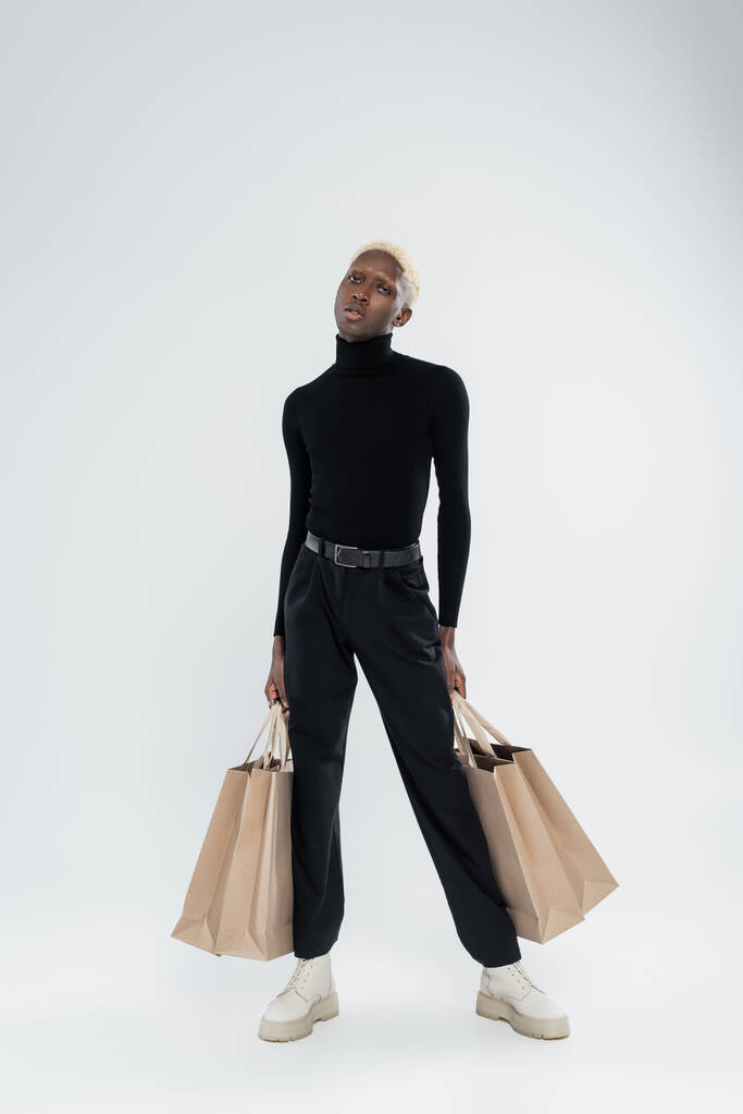plná délka blondýny africký Američan muž v úplně černé oblečení držení nákupních tašek na šedé - Fotografie, Obrázek