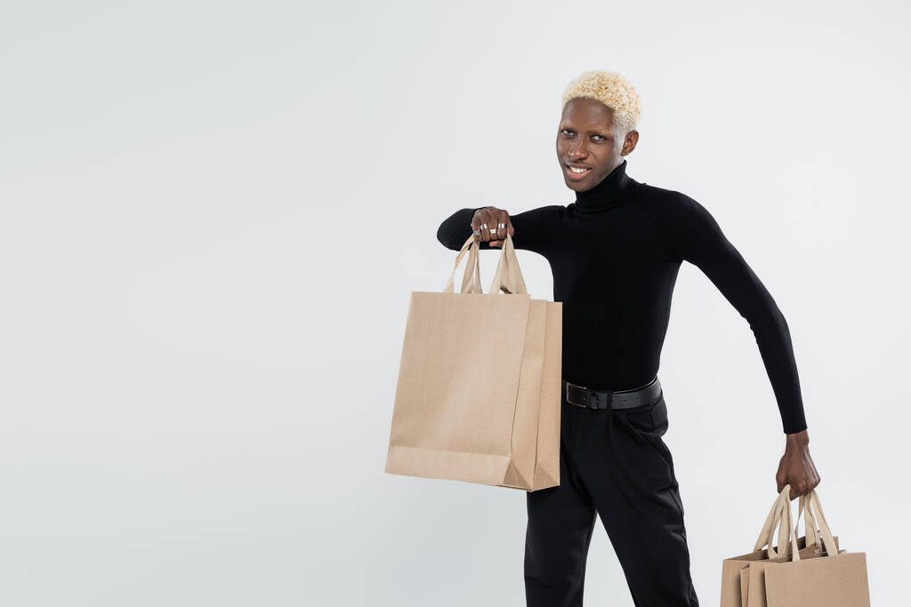 Ευτυχισμένος και ξανθός Αφροαμερικάνος με μαύρη στολή που κρατάει τσάντες για ψώνια απομονωμένες σε γκρι. - Φωτογραφία, εικόνα