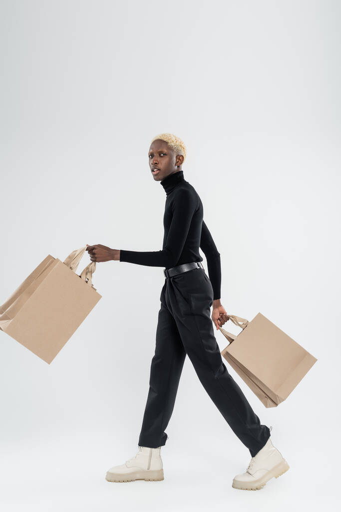 Full length of blonde African American man in totally black outfit περπάτημα με τσάντες για ψώνια σε γκρι - Φωτογραφία, εικόνα