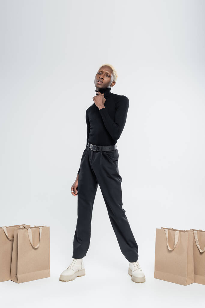 piena lunghezza di bionda afro-americana uomo in posa vicino a sacchetti di carta su grigio  - Foto, immagini