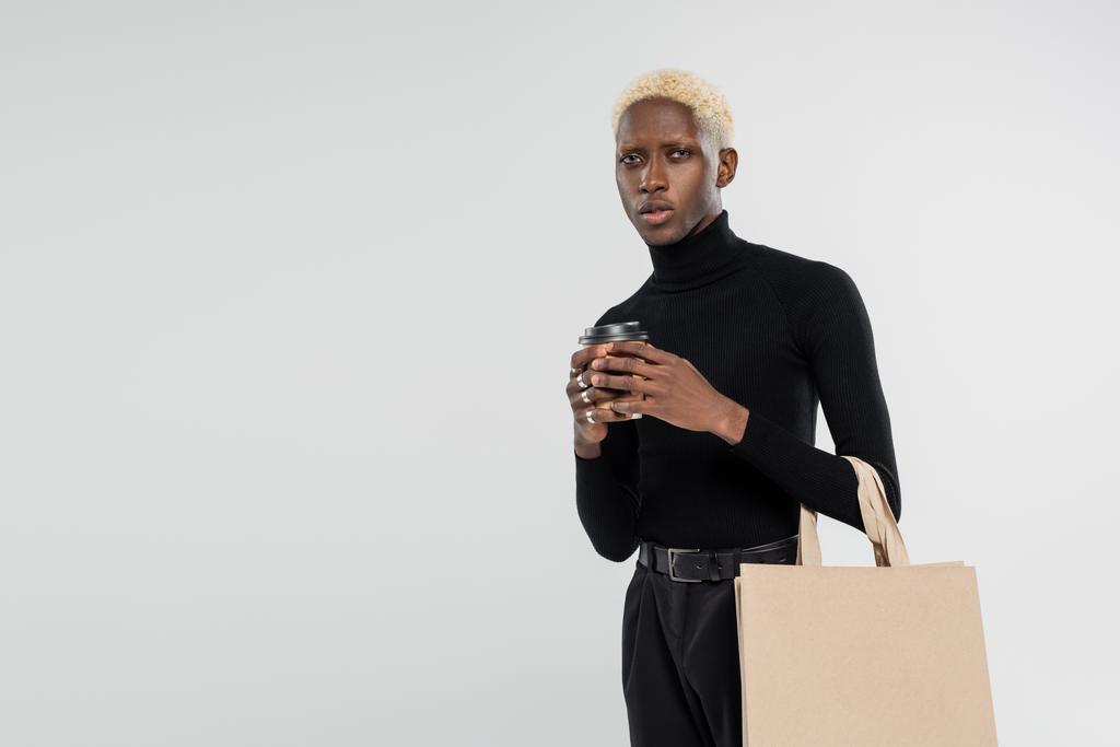 blondynka Afroamerykanin człowiek stojący z zakupy torba i papierowy kubek odizolowany na szary  - Zdjęcie, obraz