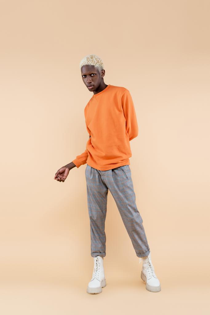 comprimento total de loira homem americano africano em camisola laranja de pé e posando em bege - Foto, Imagem