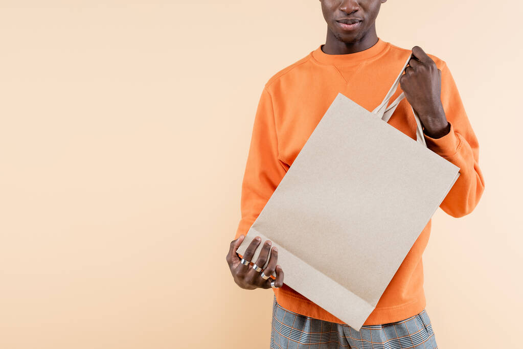 kivágott kilátás fiatal afro-amerikai férfi narancssárga pulóver gazdaság bevásárló táska elszigetelt bézs - Fotó, kép