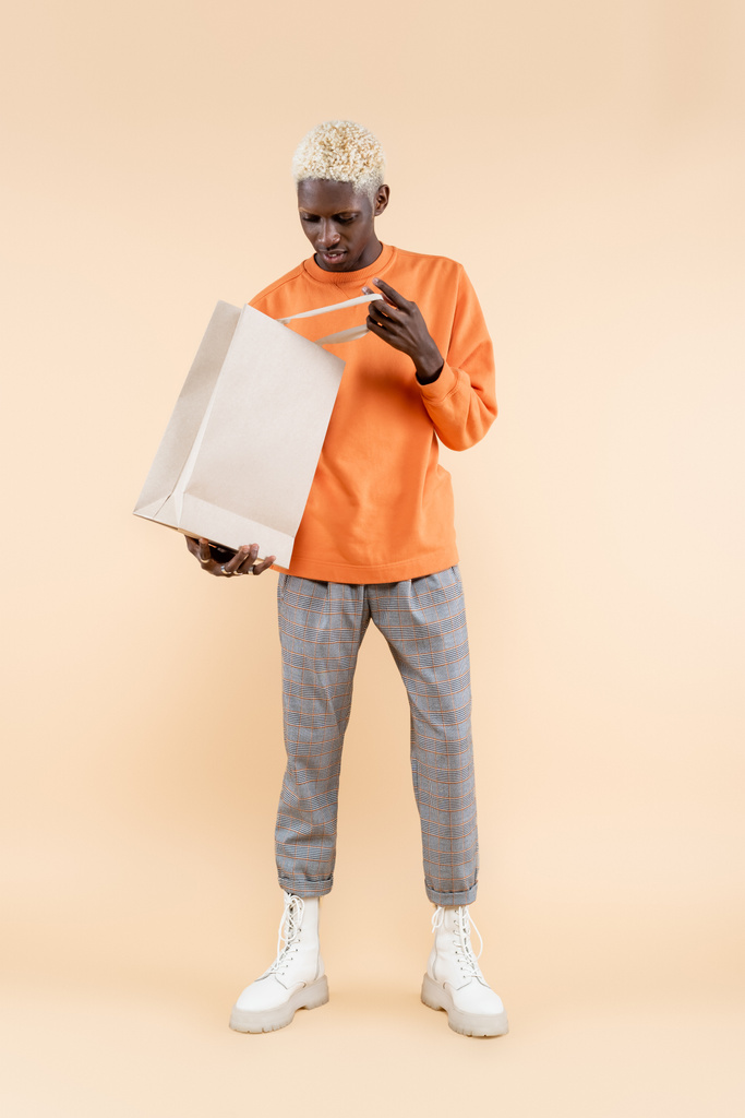 comprimento total do jovem afro-americano em camisola laranja segurando saco de compras no bege - Foto, Imagem