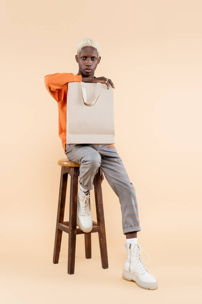 longitud completa de hombre afroamericano rubio en sudadera con bolsa de compras y sentado en la silla en beige - Foto, imagen