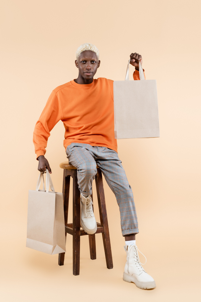 pełna długość blondynka Afroamerykanin człowiek w bluzie trzyma torby na zakupy i siedzi na krześle na beżu - Zdjęcie, obraz
