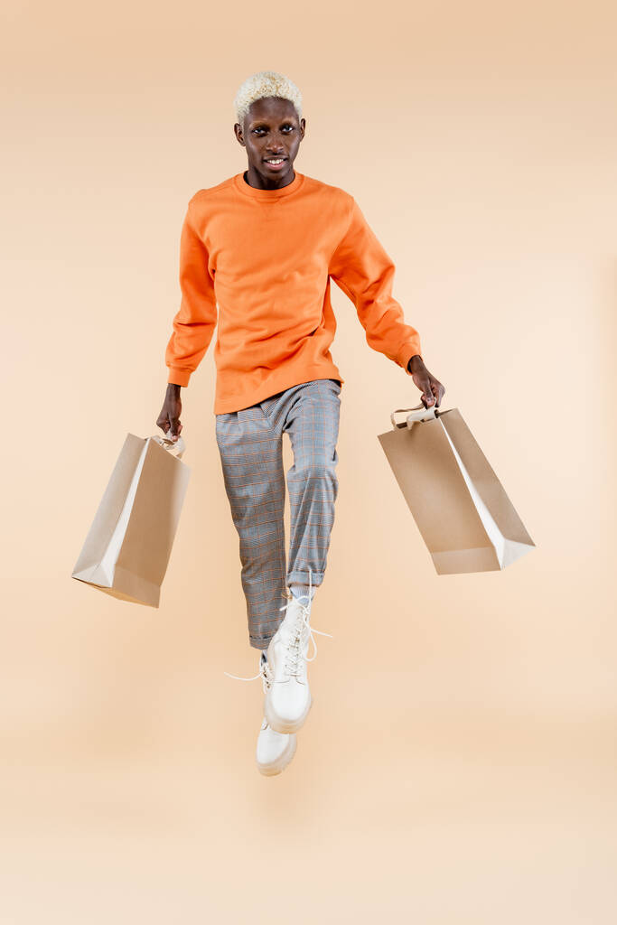 pełna długość blondynka Afroamerykanin człowiek w bluzie lewitujący i trzymając torby na zakupy na beżu - Zdjęcie, obraz