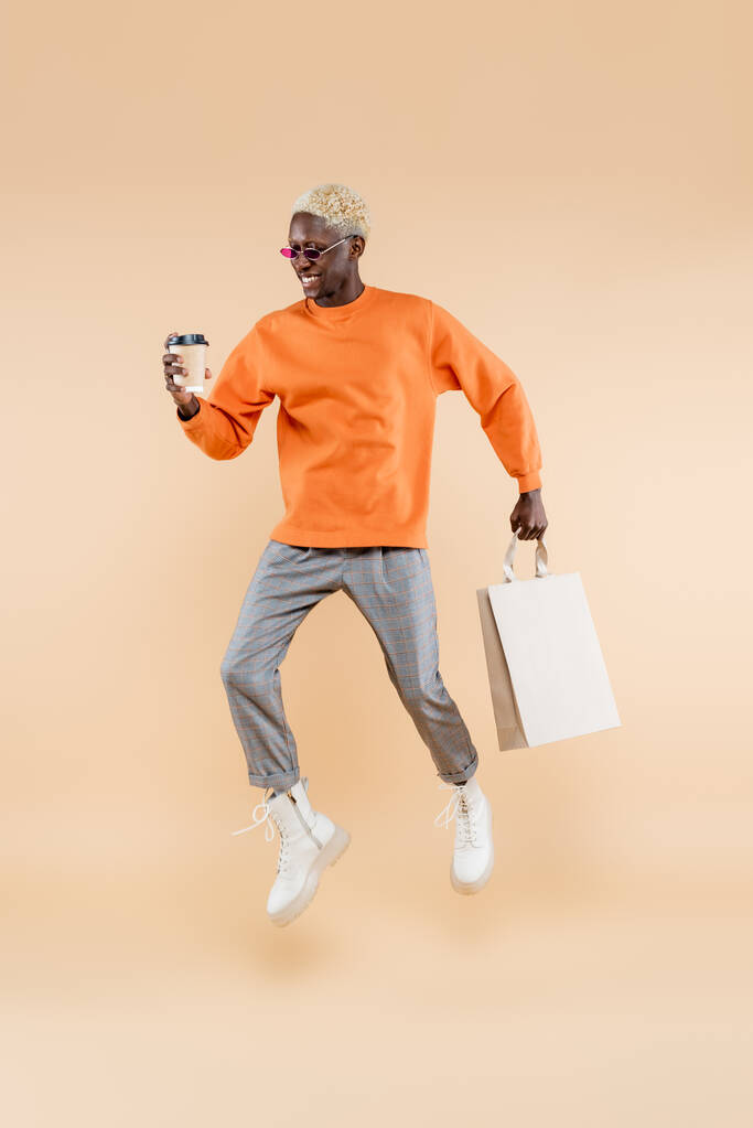 полная длина счастливый африканский американец в солнцезащитных очках левитации, держа бумажную чашку и пакет покупок на бежевый - Фото, изображение