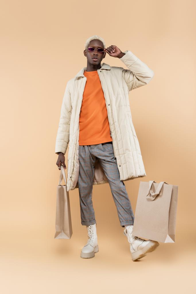 full length of trendy Αφροαμερικάνος με παλτό που προσαρμόζει γυαλιά ηλίου και κρατώντας τσάντα για ψώνια σε μπεζ - Φωτογραφία, εικόνα