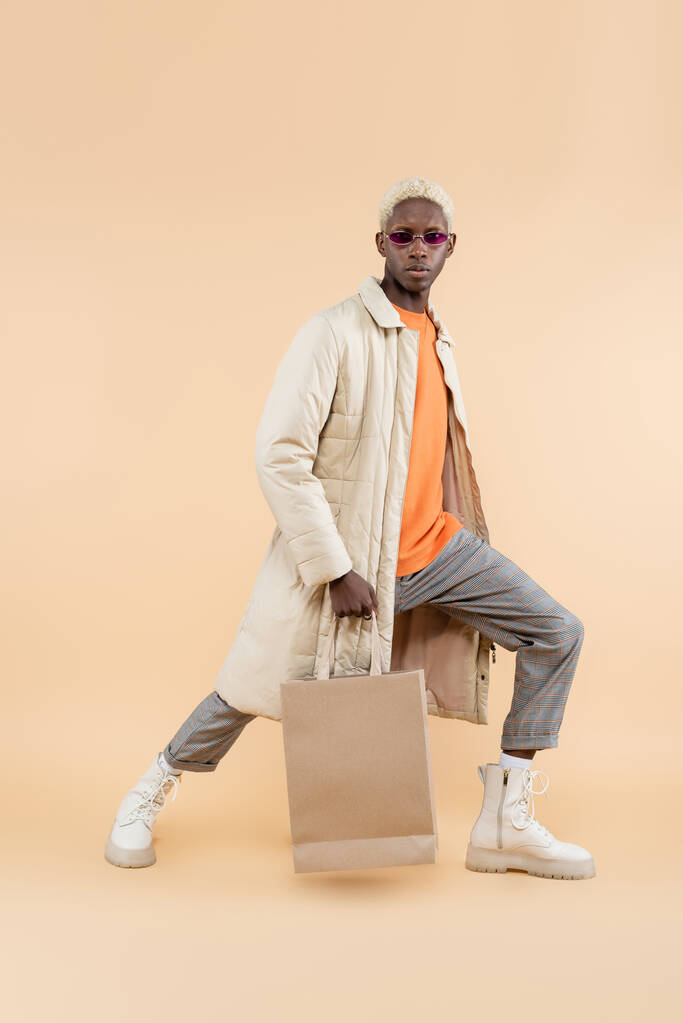コートに身を包んだアフリカ系アメリカ人男性とベージュのショッピングバッグに身を包んだサングラス - 写真・画像